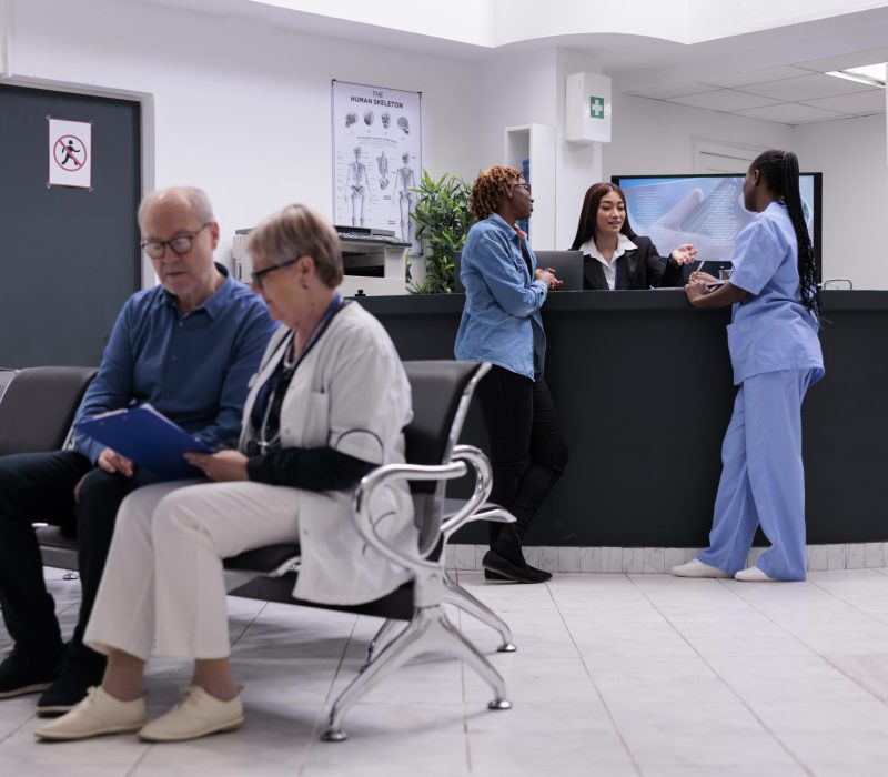 Patient and nurse sitting at reception desk, talking to female receptionist about disease diagnosis and healthcare support. Diverse people working in health center at registration counter.